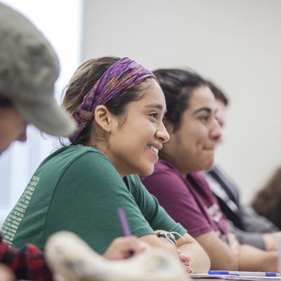 A group of smiling students attentively listening to a lecture in a classroom, with one student prominently in focus, wearing a purple headband and a green shirt.